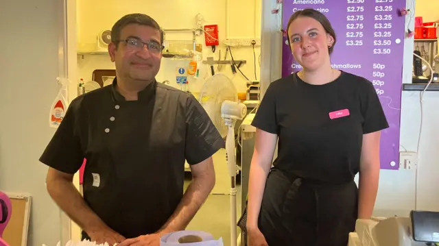 Sam Chopra wearing a black chef tunic and glasses, standing behind a counter beside a woman.