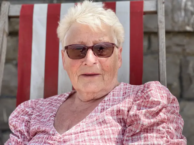 An older woman is wearing sunglasses and a red gingham blouse. She is sat in a white and red sun lounger and smiling at the camera.