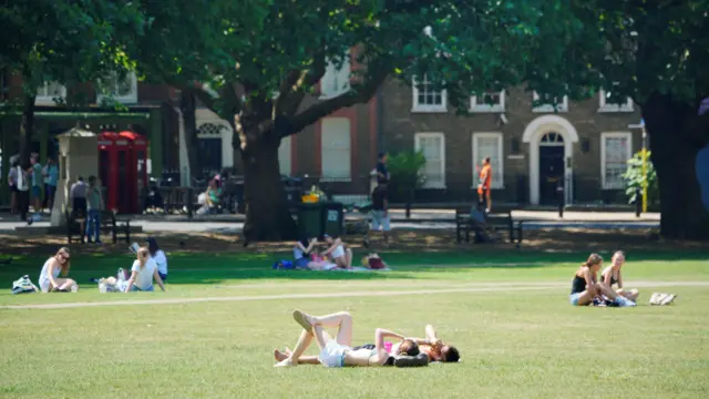 Two women cover their eyes as they lie on the grass in Richmond Green. They are surrounded by other people sat in groups enjoying the sun, while in the distance people are sat on benches and walking along the street in the shade next to a row of trees