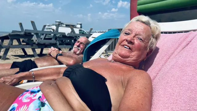A woman and a man with a white handlebar moustache are sat on sun loungers. They are tanned and red from the sun and smiling at the camera.