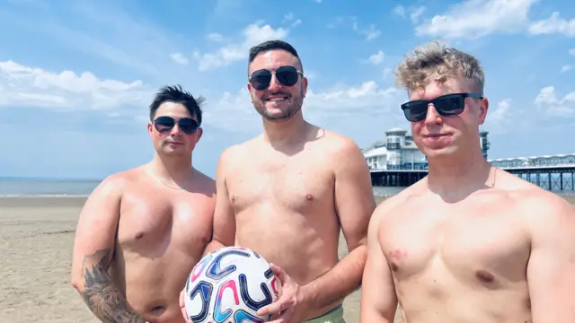 Three men are wearing sunglasses with their tops off smiling at the camera. One man in the middle is holding a football.