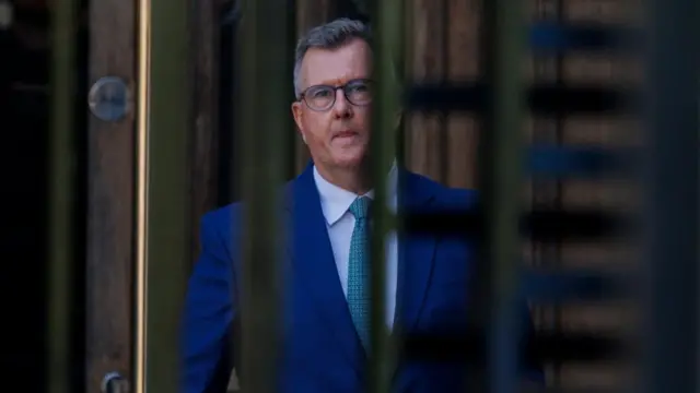 A man looks into the camera. He has grey hair and is wearing glasses. He is wearing a blue suit, white shirt and blue patterned tie. He is standing behind metal bars.