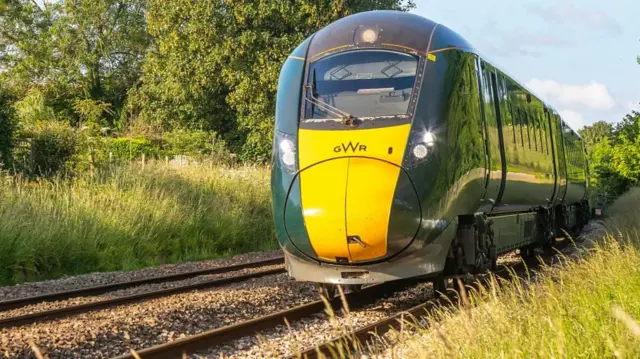 A yellow and green GWR branded train approaching the camera. It is driving along a rural stretch of the railway on a sunny day.