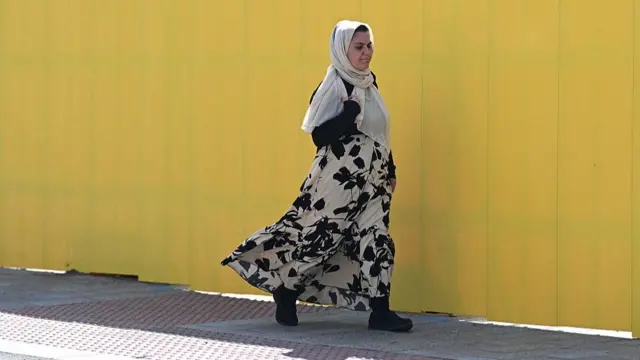 A woman in a white headscarf walks along a paved sidewalk in front of a bright yellow wall. She is walking in the shade, away from the sun