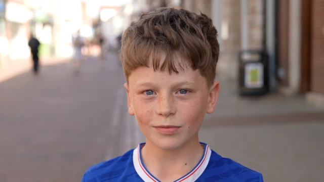 A boy smiles at the camera. He has short brown hair and wears a blue football shirt.