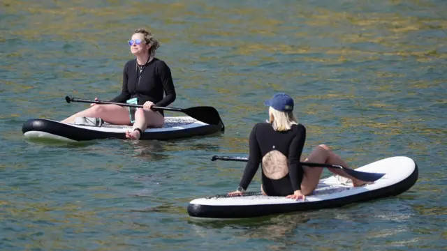 Two women sit on paddleboards in the middle of a body of water. One has her back to the camera while her oar is resting on her legs, while the other is facing the camera and looking towards the sky with sunglasses on her face