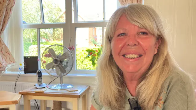Karen Macdonald wearing a light green t-shirt and smiling at the camera. She is sitting in a living room on a sunny day. Behind her is a small wooden table with a fan and landline phone on it.