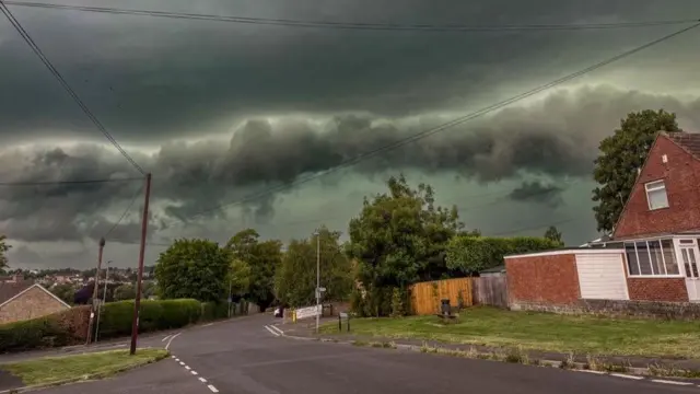A large thunderous cloud rolling towards a row of houses and trees.