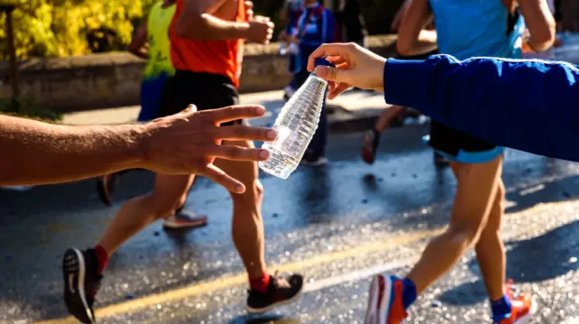 A person handing a bottle of water to a man as he runs past during a race. In the background there are other runners wearing trainers, shorts and athletic vests.