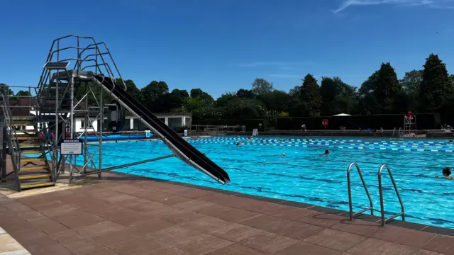 A bright blue outdoor swimming pool under a clear blue sky. There is a black slide into the pool and a ladder. There are people swimming in the pool, at the bottom end of which there is a white poolhouse. There are trees surrounding the site the lido is on.
