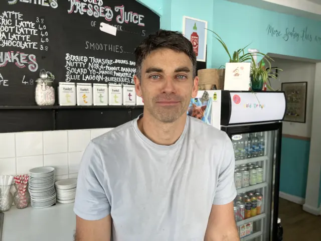 A man in a white T-shirt looks at the camera. Behind him is a cafe blackboard displaying a drinks menu.
