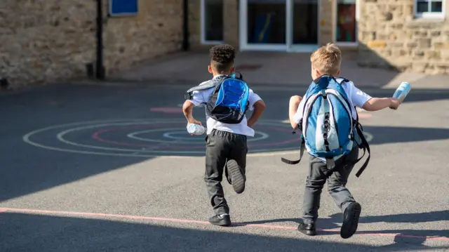 Two boys running in their school yard. They are running towards a door with their backpacks on.