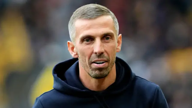 A man with short hair and a beard is pictured talking. He is wearing a navy hoodie. The background behind him is blurred.