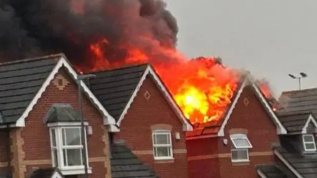 A row of detached homes, one of which is engulfed in flames coming from the roof. Black smoke is also spilling out into the air. Neighbours watch on from a distance, some of them on their phones.