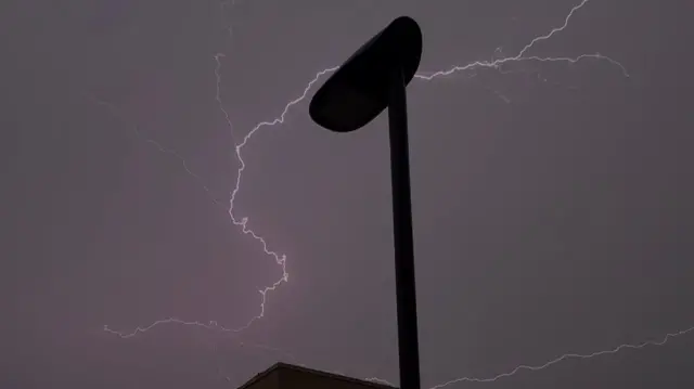 A thin purple lightning strike stretching across the night sky, against the silhouette of a lampost.
