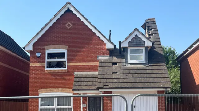 A red-brick detached home with a triangular roof and a slate awning roof. The slate section has a large hole missing.