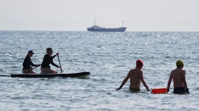 Two swimmers walk in the sea while both holding an inflatable buoy. In the distance, to their left, two people row with oars on a paddle boat, while a large ship can be seen on the horizon