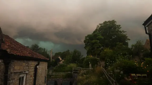 A huge dark grey cloud looming over a stone cottage and trees.