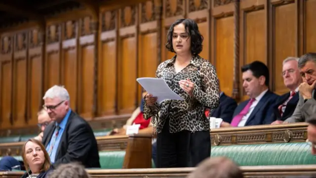 Nadia Whittome, wearing a leopard print shirt, holds pieces of paper in her hand as she gives a speech in the House of Commons. Other MPs are sitting on green benches around her in a wood-panelled room.