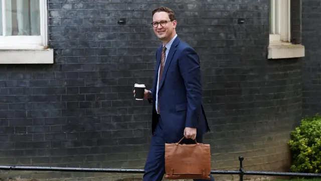 Darren Jones smiling and wearing a blue suit, holding a coffee cup in one hand and a brown bag in his left hand.