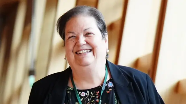 Jackie Baillie wears a dark blazer, flowery dark dress and green lanyard while walking down a corridor in the Scottish Parliament