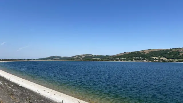 A wide shot of Cheddar Reservoir in the sun. It is a vast open water pool surrounded by a concrete border and grassy hills.
