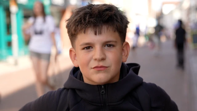 A young boy smiles at the camera as he stands outside in a High Street. He has brown hair and is wearing a black jumper.