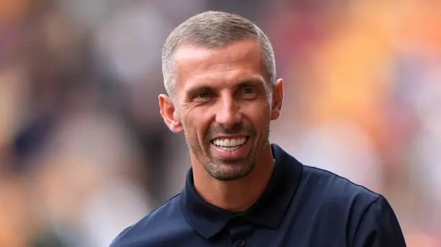 A man with short dark hair with grey in it smiles. He is wearing a dark blue polo top. The background behind him is blurred.