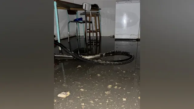 A floor is covered in debris and water. A hoover hose is lying on the floor and there are chairs in the background.