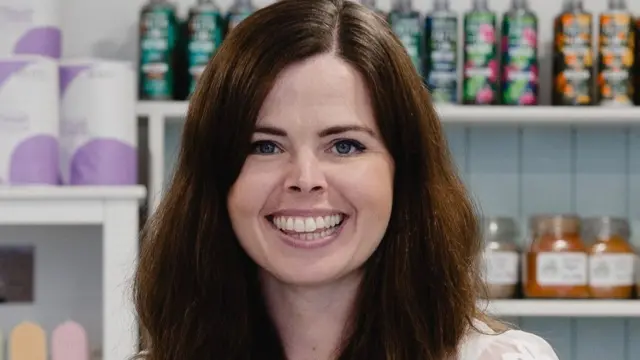 Stephanie Reed smiles at the camera while standing in a shop with jars, bottles and candles on the shelves behind her. She has long brunette hair with blue eyes and is wearing a white top with short sleeves