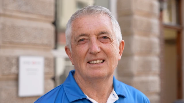 A man with grey hair smiles at the camera. He is wearing a blue polo top and is standing in front of a large brick building.