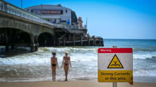 Two young women are at the edge of the sea, the pier is in the background and on the sand is a red sign that says "Strong currents in this area"