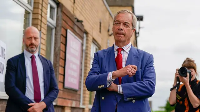Nigel Farage, wearing a blue suit, check shirt and red tie, stands outside a polling station in Makerfield during the recent by-election on 18 June. Another man in a suit, shirt and tie stands behind Farage, while a woman holding a camera up takes a photo of the Reform leader.