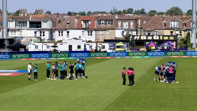 A closer shot of the cricket players on the field, standing in huddles with their respective teams.