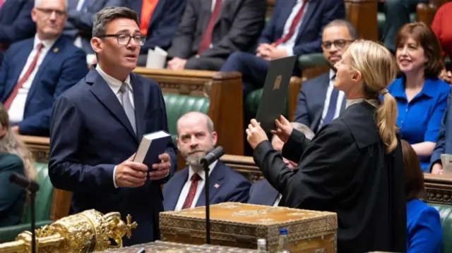 Andy Burnham is pictured at his swearing in as an MP, holding a bible in Parliament and wearing a suit and tie. A woman in front of him gestures with her hands as she directs his oath of office.