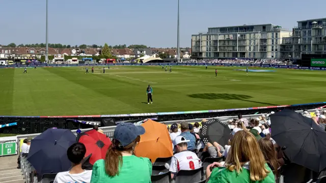 A wide shot of the players on the cricket field on a sunny day. In the foreground, spectators watch on under hats and umbrellas.