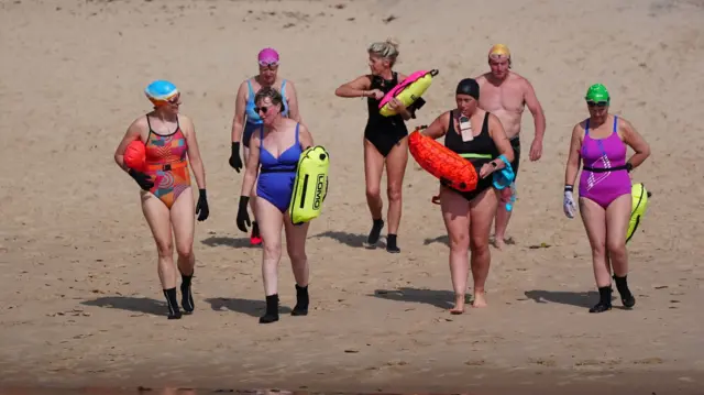 A group of swimmers are walking towards the water's edge on a sandy beach. Six women are wearing bathing suits in a range of colours and one man is wearing a gold swim cap and black trunks