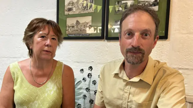 Two people, a man and a woman, sat next to each other looking into the camera.