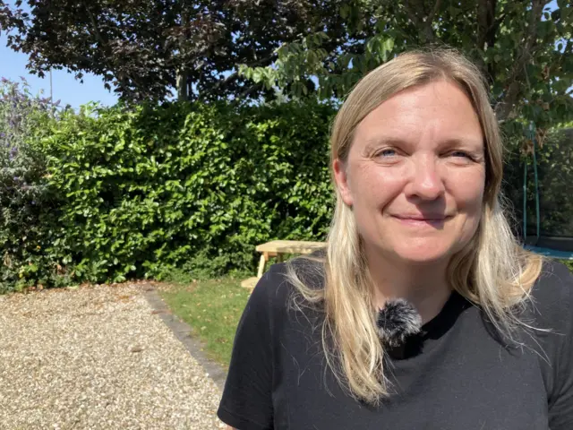 Laura, who has blonde hair and wears a black t-shirt, looks at the camera with a worried smile.