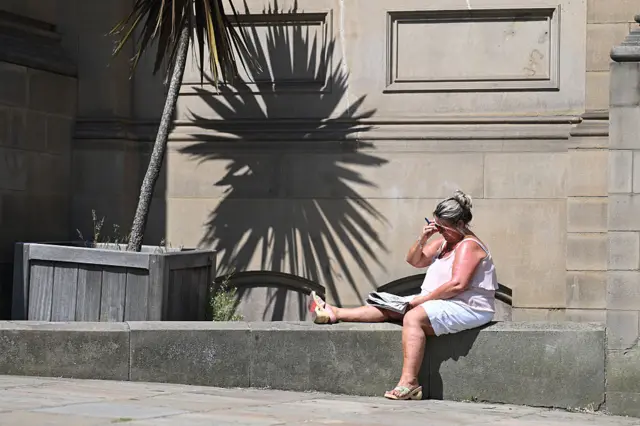 A lady sits on a ledge with her leg up as she reads the paper as the sun shines on her in Huddersfield
