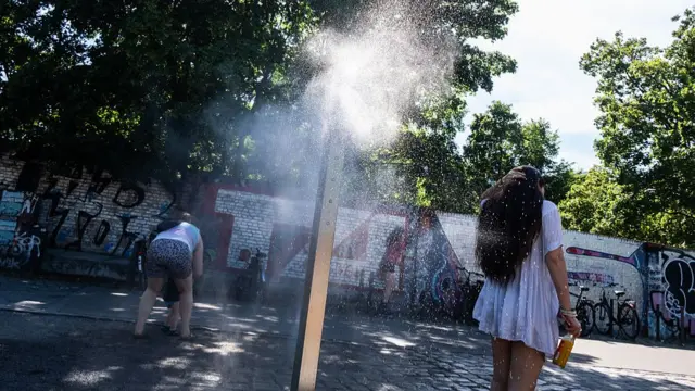 A woman strokes her hair while she stands next to a mist spraying fountain in the middle of a stone paved street, with a wall of graffiti in the background. To her left is a woman in leopard print shorts and white top tending to her child