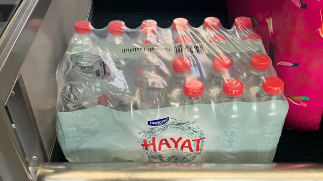 Water bottles with a red cap are positioned in the luggage rack, with a pink suitcase to the right and a grey camo one beneath the bottles