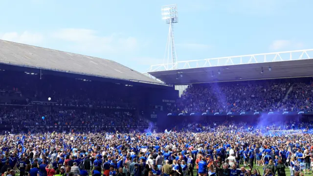 Fans storm a football ground within a stadium in celebration after Ipswich Town was promoted back up to the Premier League. Fans are dressed in blue football shirts and some blue flares billow into the sky on a sunny day.