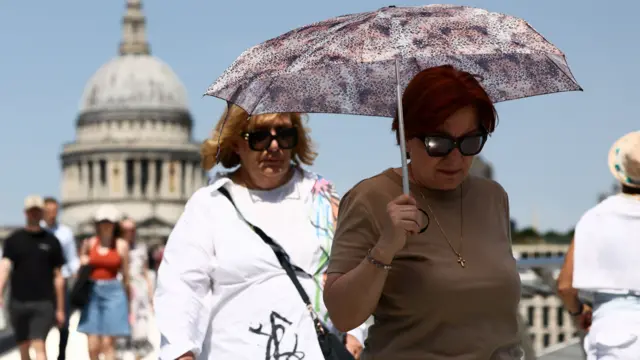 A woman wearing sunglasses walks with an umbrella along the Millennium Bridge in London. Another woman, also wearing sunglasses, is walking close behind her