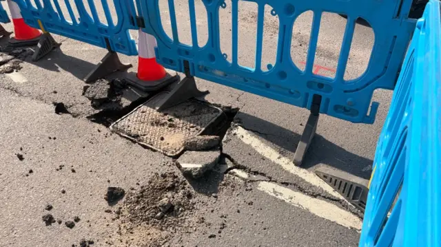 A close up shot of a manhole cover on a road, with jagged chunks of asphalt. It is surrounded by blue plastic barriers and orange traffic cones.