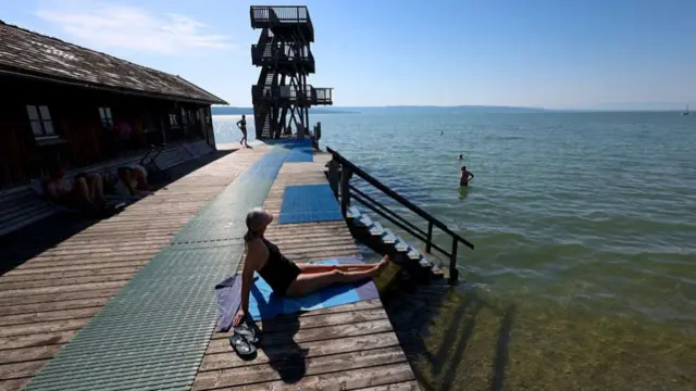A woman in a black bathing suits and sun cap sits on a wooden jetty next to a wooden diving tower at a lake where other people are also swimming