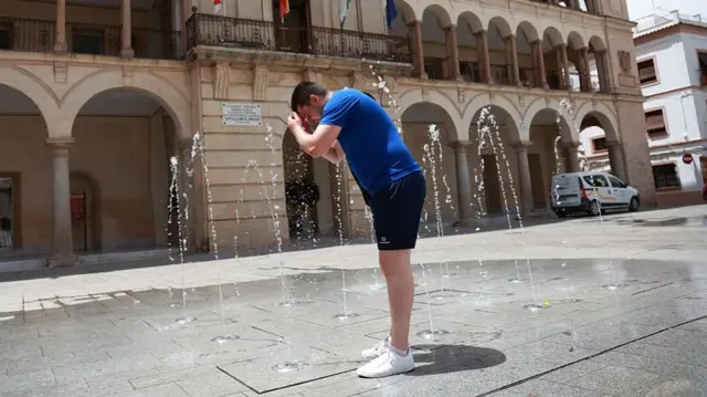 A man wearing a blue top, black shorts and white trainers is standing in a public square, cooling himself off with water that is spraying from the ground