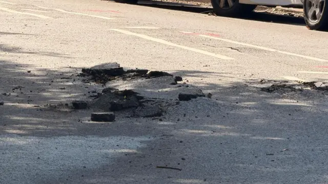 The site where a lightning strike has fractured the surface of the road, leaving a shallow crater with jagged and uprooted edges.