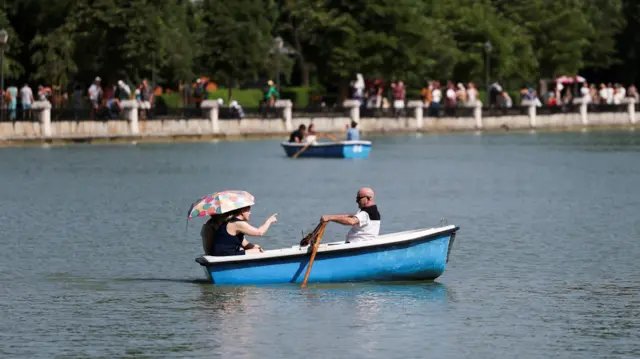 A bald man with sunglasses rows a blue dingy in a lake while two other people hide under an umbrella at the back of the boat. Another boat can be seen in distance, while crowds of people walk along the waters edge