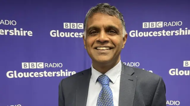 Dr Anathakrishnan Raghuram smiles in front of a wall branded with BBC Radio Gloucestershire. He has dark grey hair and wears a grey suit with a light shirt and blue tie.